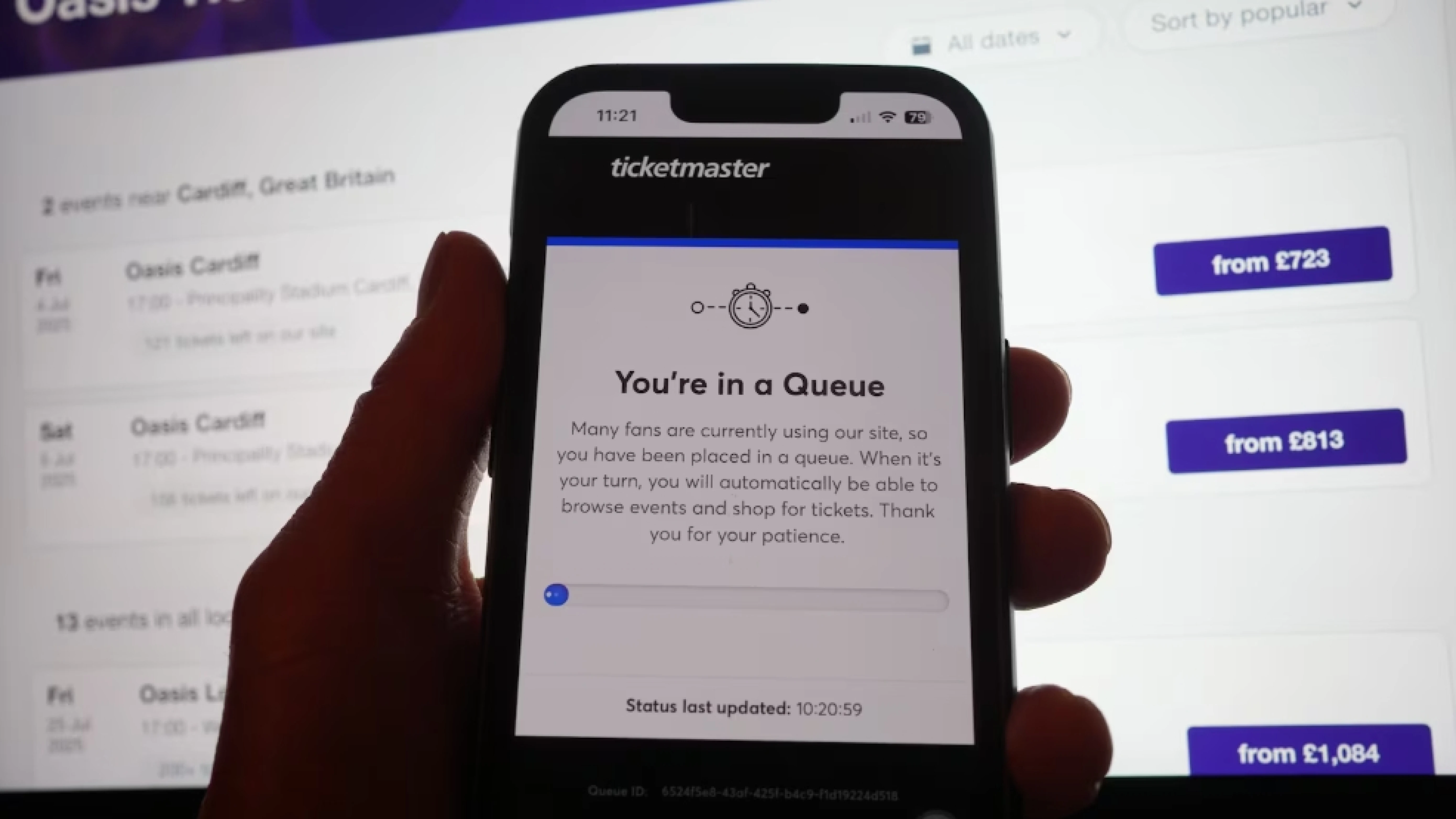 Close-up picture of smartphone being held in front of a computer display showing Oasis Ticketmaster prices. The closer phone screen reads: You're in a queue. Credit: Yui Mok / PA